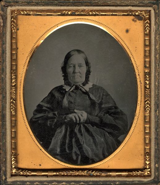 Sixth plate ferrotype/tintype of an unidentified woman, described as the mother of Alexander Van Pelt. Waist-up portrait, facing forward, with her hands across waist. She is wearing a dark dress, white collar, and dark tied bonnet. Hand-coloring on cheeks. 