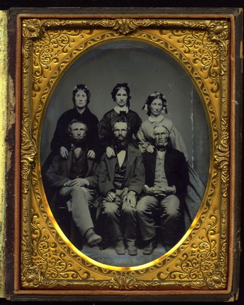 Half plate ferrotype/tintype of three men and three women. The men are seated in front, wearing suits, with their hands in their laps. The man on the left has crossed legs. Behind them are standing three women, all wearing bonnets with ribbons, and their hands resting on the men's shoulders. Hand-coloring on cheeks. 