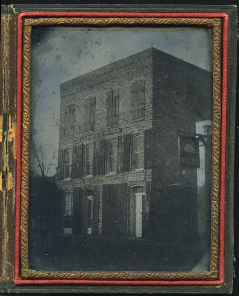 Quarter plate ambrotype of a brick structure, possibly in Green Bay, Wisconsin, housing the office of J.S. Baker, Land Agent. The building is three stories, with four bays on each story, and six-over-six double-hung windows. Shutters on top story are closed. Doorway is on far right. On the right is the New England House, c. 1855-1865.