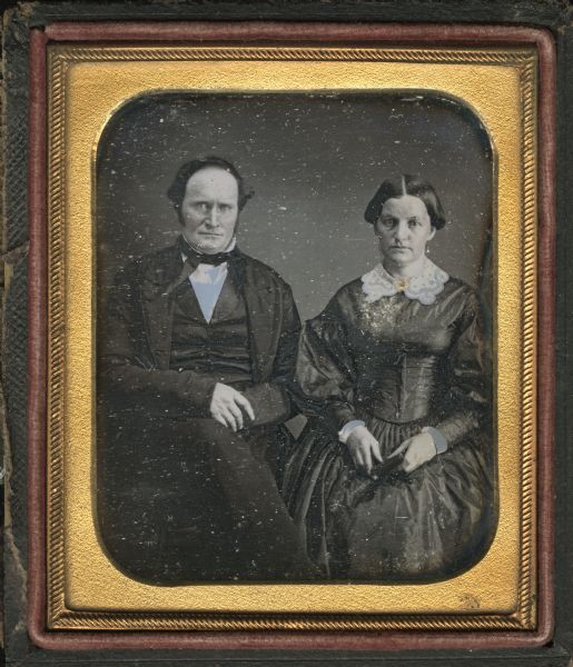 Sixth plate daguerreotype of James Harvey Sanford and Rebecca Johnson Sanford. Three-quarter length portrait, both seated, facing forward, with hands in laps. James is wearing a suit, tie, and stand collar, and his legs are crossed. Rebecca is wearing a dark dress with balloon sleeves, white collar and cuffs, and gold brooch at her neck, and is holding a small book or a cased photograph. Hand-coloring on cheeks. 