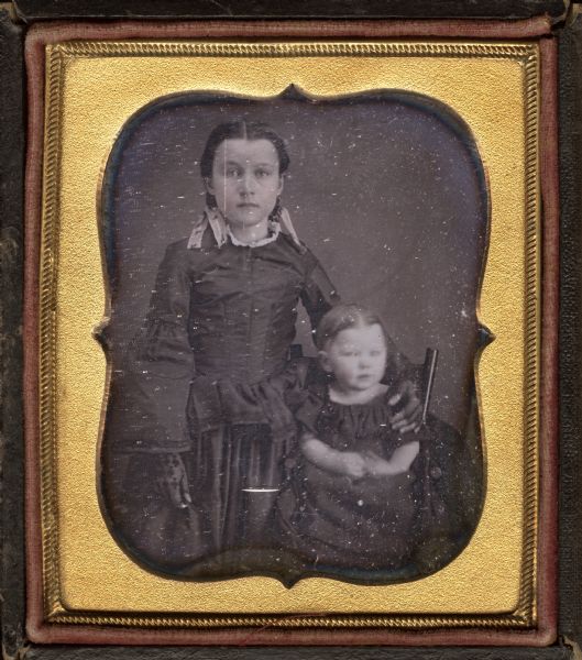 Sixth plate daguerreotype of Mary and Nancy Sanford as children. The older child is standing with her left arm around the shoulder of the toddler, who is sitting on a high chair, and is wearing a dark dress with peplum, bell sleeves, and lace mitts, and ribbons in her hair. The toddler is wearing a dark dress with heavy gathers at the neck. Hand-coloring on cheeks. 