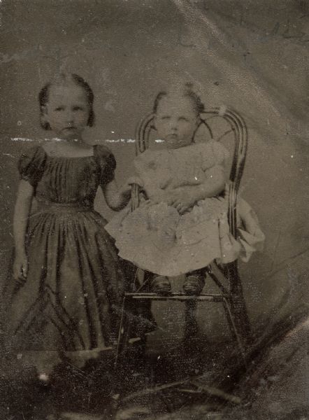 Sixth plate ferrotype/tintype of Nancy Sanford and an unidentified girl. Full-length portrait, facing front. The elder girl is standing with her hand on the arm of the wicker high chair on which the toddler is sitting, and is wearing a square-yoked, short sleeved, heavily gathered dark dress belted at the waist. The toddler is in a light-colored, short sleeved dress with ruffled skirt. Hand-coloring on cheeks. 