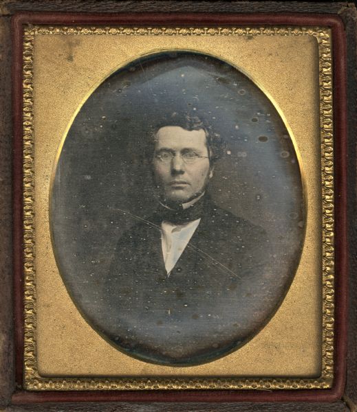 Sixth plate daguerreotype of a member of the George A. Johnson family. Waist-up portrait facing forward. He is wearing a suit, cravat, stand collar, and octagonal spectacles. Hand-coloring on cheeks and lips. 