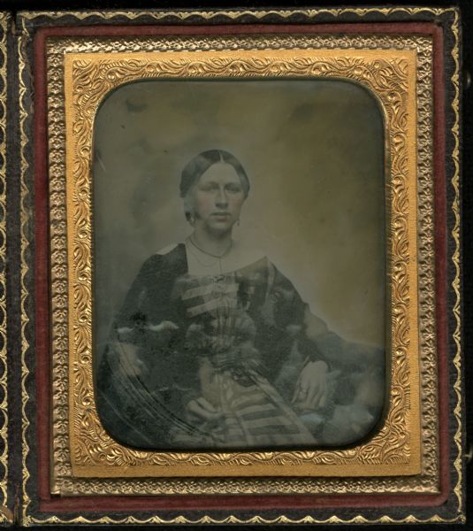 Sixth plate ambrotype of an unidentified member of the George A. Johnson family. Waist-up portrait. She is sitting, facing forward, resting her arm on unidentified object, with her hand in her lap. She is wearing a striped, off-the-shoulder dress, a ring on her forefinger, drop earrings, and a chain necklace. Hand-coloring on cheeks and lips. 
