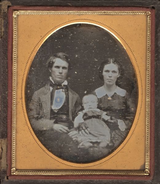 Sixth plate daguerreotype of an unidentified couple with a sleeping infant. Waist-up portrait, seated, facing forward. He is wearing a suit with light-colored jacket, stand collar, and long bow tie, she is wearing a dark dress, white cutwork collar, mitts, and gold brooch. Hand-coloring on cheeks and brooch. 