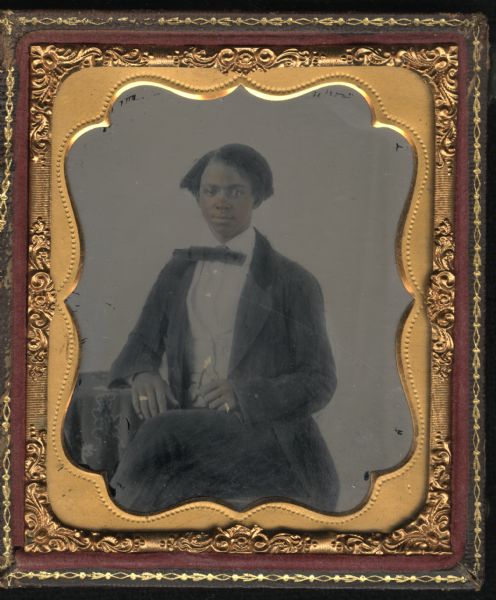 Sixth plate ambrotype of an unidentified African-American man. Three-quarter length portrait, seated, facing forward, with torso turned slightly left, legs crossed, and forearm resting on a cloth-covered table. He is wearing a suit with velvet collar, a white vest, shirt collar turned down, and narrow horizontal bow tie. A watch chain hangs from his vest, and a gold ring is on each hand. Hand-coloring on cheeks and gold details on jewelry. 