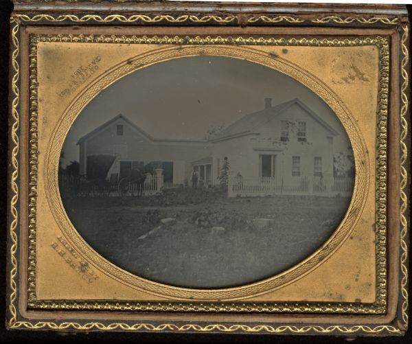 Quarter plate ambrotype of a view of a two-story white house, with its gable end facing forward, front door and two windows on the first floor, and two windows on the second. A one-story, porticoed addition connects it to the barn or stable, which has two open doors. A picket fence, covered buggy with one horse in harness, and a man holding the hands of two children are in front of the house. 