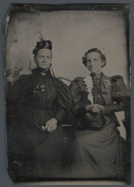 Ferrotype/tintype of M.E. Stewart and Mrs. I.B. Woodward. Three-quarter length portrait, both seated, facing forward, in front of a painted backdrop. They are wearing dark dresses with balloon sleeves, collar pins, and flowers pinned on the front of their dresses. The woman on the left is wearing a tied bonnet with feathers, and is holding a small bag. The woman on the right is wearing gloves and is holding her hat.