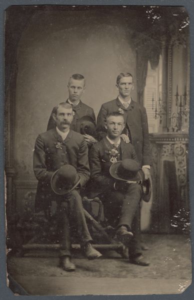 Ferrotype/tintype portrait of three students with their principal, taken in a studio in front of a painted backdrop, the day after their high school graduation. The principal, William F. Gray, is seated on the left, next to William D. Lamont. Standing are Henry Chappel on the left, and David E. Kiser on the right. The men are wearing suits and floral boutonnieres, and are holding their hats. 