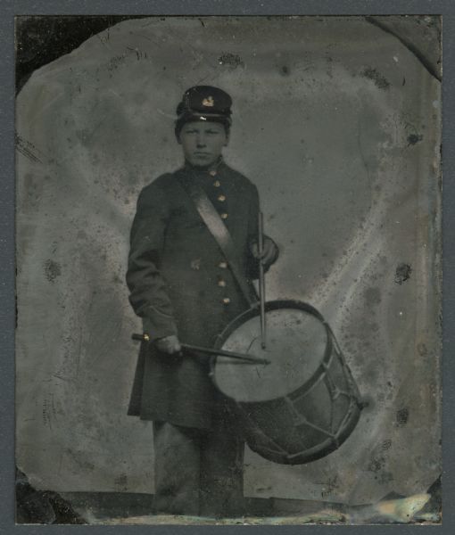 Sixth plate ferrotype/tintype of Ceylon C. Lincoln as a young drummer boy in the Union Army. Three-quarter length portrait, standing, facing forward. He is wearing his military uniform and cap, and is holding drumsticks over his drum, which is strapped over his shoulder. Hand-coloring on cheeks and gold on uniform buttons.