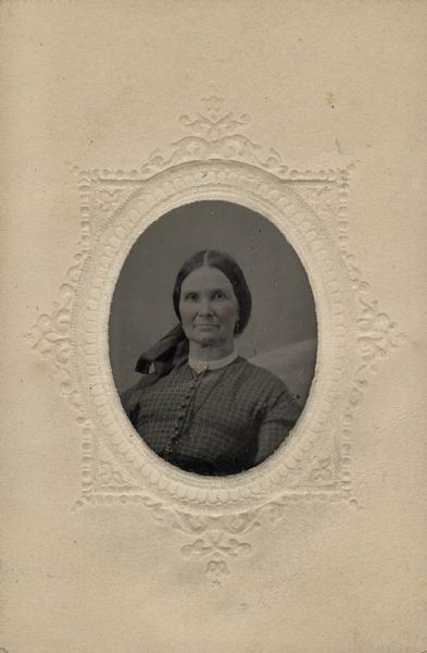 Sixteenth plate ferrotype/tintype of Lucinda Alice Putnam Perkins (1812-1910). Quarter-length portrait, facing forward. She is wearing a print dress buttoned down the front, a white collar, and a brooch at the neck. Her hair is tied back with a long ribbon. Hand-coloring on cheeks.