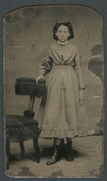 Ferrotype/tintype of Susanah Arnold Frazee. Full-length portrait, standing, facing forward, with hand holding the back of a chair. She is wearing a belted dress with ruffled hem and white collar, a brooch at her neck, and high-top button-up shoes. Hand-coloring on cheeks, and gold detail on brooch. The posing stand is visible behind her shoes and at her left shoulder. 