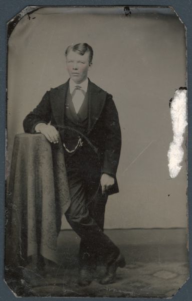 Ferrotype/tintype of a young Frank Miller. Full-length portrait, standing, facing forward, with one arm resting on a cloth-covered stand, with one leg crossed over the other. He is wearing a suit coat with very wide lapels, and a watch chain from his vest. Hand-coloring on cheeks. 