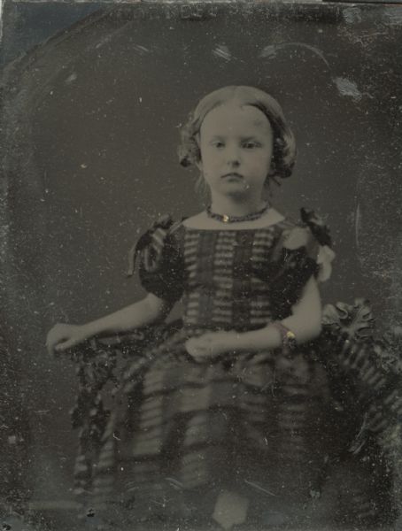 Ferrotype/tintype of Minnie Beers, daughter of William and Agnes Beers. She is sitting and resting her arm on the arm of a cast iron chair. She is wearing a short puffed sleeve striped dress with bows at the shoulders, a beaded choker and gold bracelets. Hand-coloring on cheeks and gold details on jewelry. 