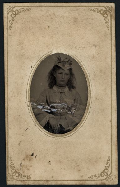 Ferrotype/tintype of Anna McIntyre. Waist-up portrait facing forward. She is wearing a light-colored dress with fringed cuffs, a bow at the neck, two pendant necklaces, a choker, two rings on her right hand, and a hat decorated with flower sprigs. Her shoulder-length hair is loose and uncombed. Hand-coloring on cheeks.