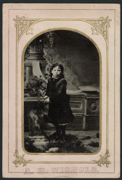 Ferrotype/tintype of a full-length studio portrait of Clara Demarel standing in front of a painted backdrop. She is leaning on a prop pillar with a potted plant on top, and wears a knee-length dark coat and a cross pendant. Cheeks hand-colored. 