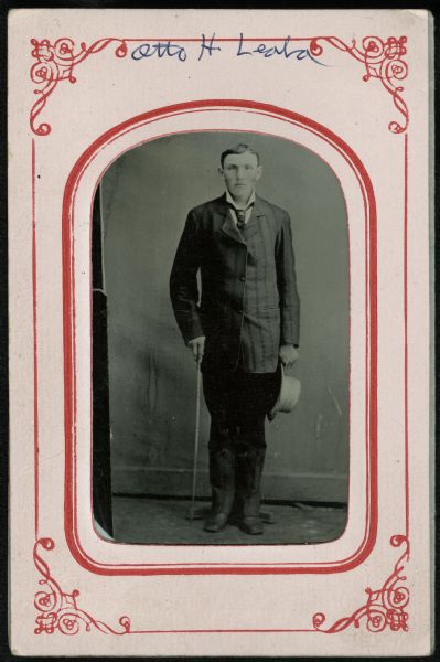 Ferrotype/tintype of a full-length portrait of Otto H. Leda standing. He is wearing a suit and necktie, and is holding a cane in one hand, and a hat in the other. Hand-coloring on cheeks.