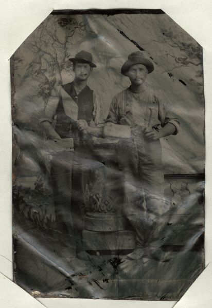 Ferrotype/tintype of a full-length portrait of two men posing with bricks and trowels in front of a painted backdrop. They are standing next to a faux stone balustrade. One of the men appears to have a cigar in his mouth.