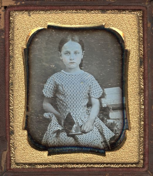 Sixth plate daguerreotype of a three-quarter length portrait of an unidentified girl wearing a short sleeved striped dress and holding a cased image in her lap. She is facing forward and is seated next to a cloth-covered table. On the table are books and covered boxes. The cased image appears to be that of a child. Hand-coloring on lips and cheeks. 
