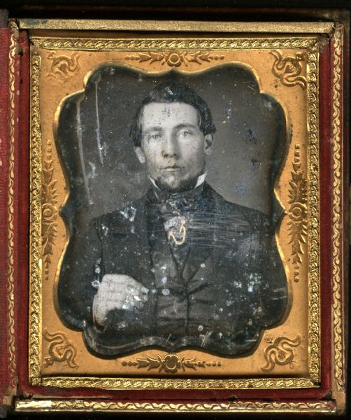 Sixth plate daguerreotype of an unidentified man. Waist-up portrait, facing forward, he is wearing a suit, cravat, stand collar, and a watch chain attached to his vest, and is holding his lapel. Hand-coloring on cheeks and chain. 
