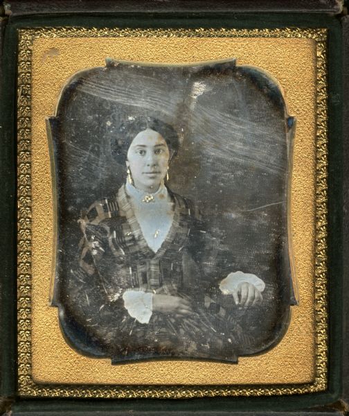 Sixth plate daguerreotype of an unidentified woman. Waist-up portrait, seated, facing forward, with her arm on a prop. She is wearing a plaid dress in the habit front style over a white chemise with white cutwork sleeves. Hand-coloring on cheeks, drop earrings, and a brooch at the neck.