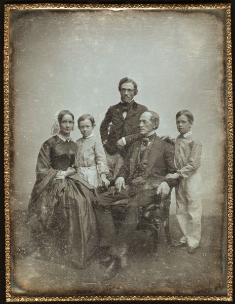 Whole plate daguerreotype of the Allcott family. The parents, Sidney S. and his wife Julia, are seated. Sidney faces partially left, Julia forward. Their three sons are standing, facing forward, and are, left to right, Frank, George, and Charles. The two younger sons are wearing identical white pants and checked shirts. Sidney and George are wearing suits and cravats. George is holding a cane in his left hand. Julia is in a dark dress, white lace collar, white cap, and fringed shawl. 
George first went to California in 1849. While on a return trip there, he died at Acapulci, Mexico. 