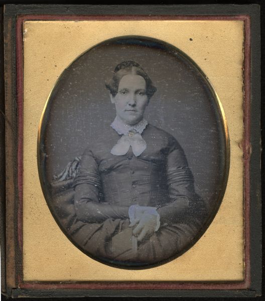 Sixth plate daguerreotype of a waist-up portrait of an unidentified woman. She is seated on an upholstered chair with carved top, facing front. Her hands are in her lap holding a book. She is wearing a dark dress, white embroidered collar, and ribbon crossed at the neck and held with a brooch. Hand-coloring on cheeks, lips, and brooch.