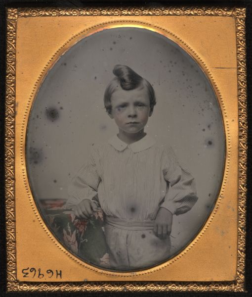 Sixth plate ambrotype of an unidentified boy. Three-quarter length portrait, standing facing forward, with his arm resting on a cloth covered table. He is wearing white trousers, a striped white shirt with white collar, and a topknot hairstyle. Hand-coloring on cheeks and table cloth.