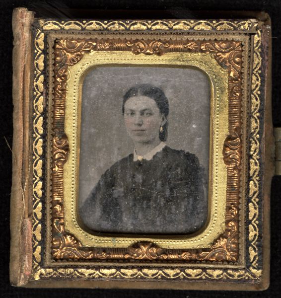 Sixteenth plate ferrotype/tintype of a quarter-length portrait of an unidentified woman. She is facing front and slightly left, and is wearing a dark dress, white collar, gold brooch at the neck, and gold drop earrings. Hand-coloring on cheeks, lips, and jewelry. 