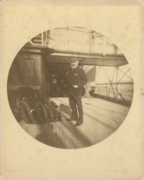 Frederick Layton standing on deck smoking a cigar while at sea in a Kodak #2 camera portrait by T.W. Ingersoll from St. Paul, Minnesota. The photographer's shadow is in the foreground. There is a second man sitting in a deck chair on the left, covered with a blanket.