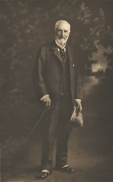 Full-length portrait in front of a painted backdrop of Frederick Layton in late middle age. He is holding a cane in one hand and a hat in the other. He died in 1919.