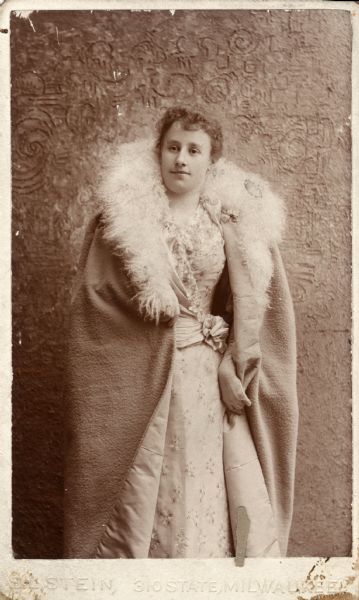 Three-quarter length portrait of Grace Hayman Fowle. She is wearing gloves, and a fur-trimmed cloak over a flower printed and ruffled dress. Grace was the niece of Elizabeth Hayman Layton.