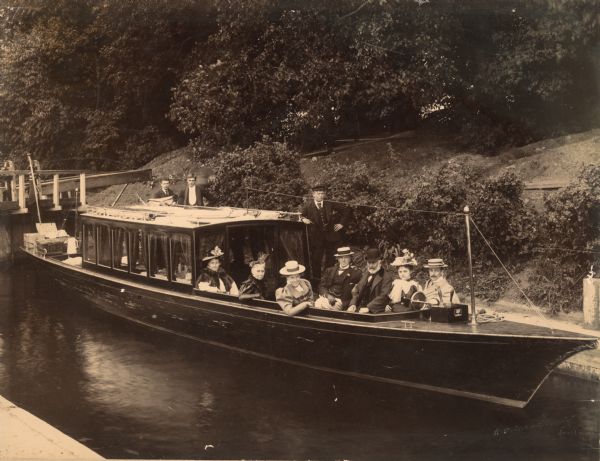 View from shoreline of excursion boat docked in a wooded area. From left to right: Mary ??, Elizabeth Layton, Grace Hayman, Henry Wallis (owner of the French Gallery, Pall Mall), Frederick Layton, Laura Ackroyd (poet), and the son of Henry Wallis. There are three boatmen, including the captain, standing behind the boat cabin. On the far left at the end of the boat are picnic baskets.