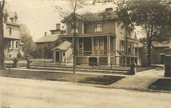 A view across street towards Frederick and Elizabeth Layton's home at 524 Marshall Street. The clapboard house has two porches. On the left is a roofed enclosure covering the front steps. Folded awnings are on the windows. The property is fronted by a cement and iron fence. Below the fence, the terrace is edged with stones. They lived in this house all their married life. Elizabeth died in 1910; Frederick in 1919.