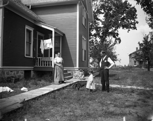 Exterior view of family posing on the lawn in front of a house. The woman on the left is standing on a board sidewalk holding a broom. Behind her, another woman sits on the sidewalk washing a baby in a wash bowl. A man, a small child and a dog are standing in the grass. Railroad tracks are in the background.