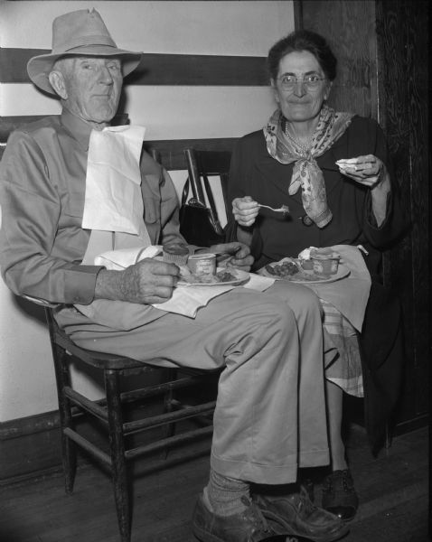Interior portrait of a farmer and his wife sitting on chairs and eating off paper plates in their laps. There are containers of "Meadow Gold" on their plates.