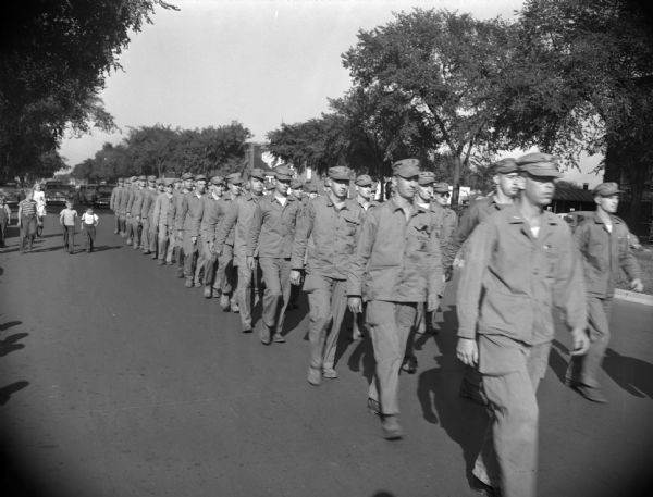 A platoon of Marines marching down a tree-lined city street. A group of children are walking alongside on the left.