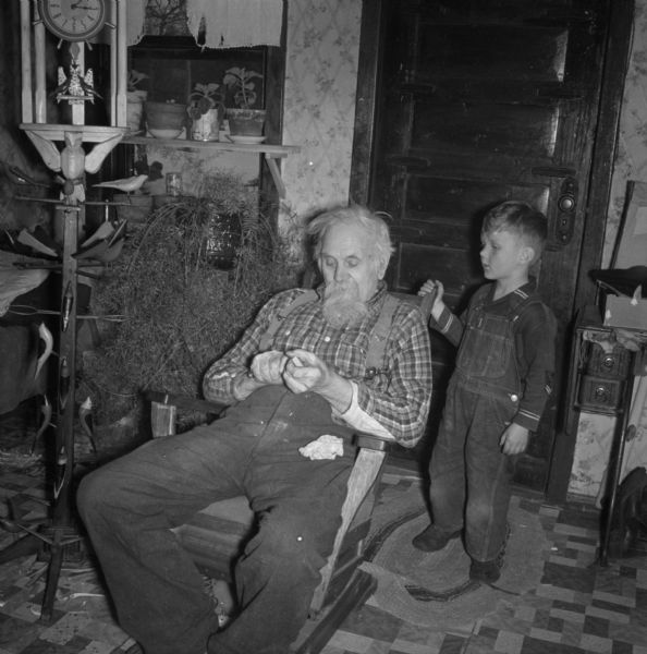 Interior portrait of an elderly man sitting in a rocking chair whittling. A small boy is standing behind him on the right. Potted plants are on the window sill in the background. On the left is a clock on a stand decorated with wooden birds.