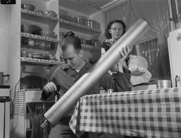 Interior portrait of a man sitting at a kitchen table adjusting his homemade telescope. His wife is washing dishes behind him. Open shelves on the wall display pots and dishes.