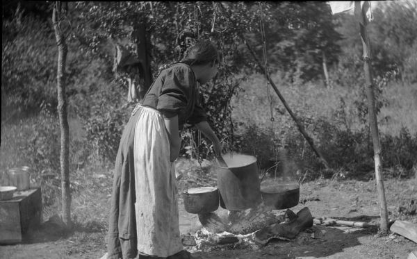 Chippewa woman standing outdoors stirring one of three pots suspended over an open fire.
