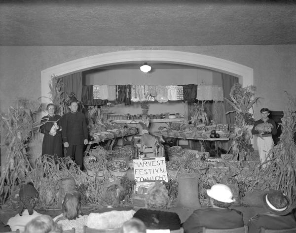 Interior view, over the heads of an audience sitting in the foreground, of a room filled with vegetable produce. A man, and a woman holding a puppy, are standing on the left wearing Salvation Army uniforms. A boy standing on the right is holding a large rabbit. A goat and a puppy sit in a cart in the center of the display. Clothes hanging from a line along the back wall, above preserved food in jars displayed on a table,