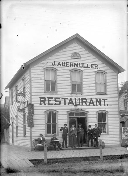 Exterior view from unpaved street of a group of men enjoying mugs of beer while standing at the front entrance of J. Auermuller Restaurant and Saloon. Two other men are sitting on a bench on the left.