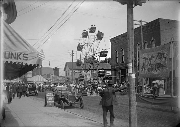 View from sidewalk of a street carnival with tents, circus banners and a Ferris Wheel set up in the middle of the street. Signboards advertise ice cream and postcard photographs, and automobiles are parked along the curb near storefronts.