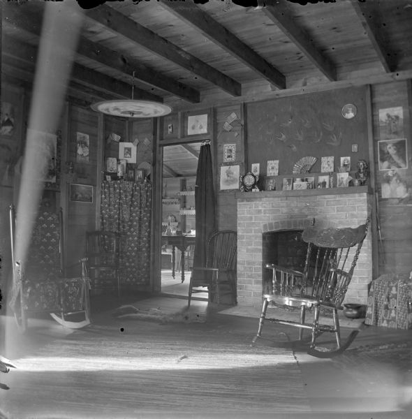 Interior of a room with exposed walls and ceilings. Straight backed chairs and rocking chairs are in the room near a fireplace on the right. Assorted photographs decorate the walls, and more photographs, prints, and a clock are on the mantel. An open umbrella hangs from one of the the ceiling rafters on the left. An open doorway in the background on the left leads to a room with a table and chair, with tableware on open shelves on the back wall.