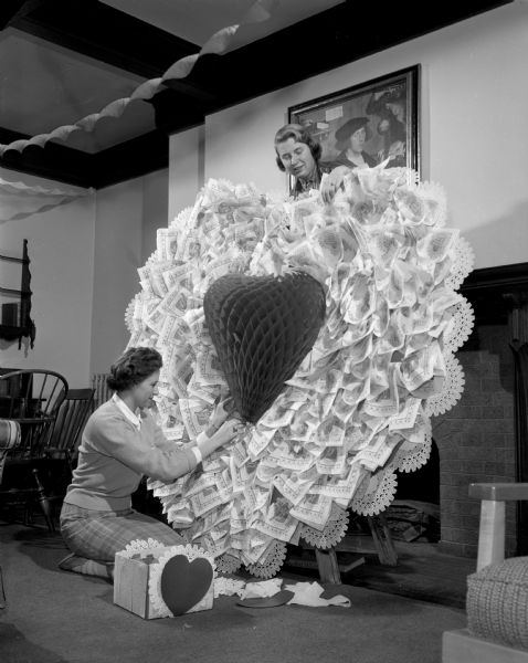Two women are assembling a large paper valentine decoration. One woman stands on a ladder in front of a fireplace working on the top of the decoration, while another woman kneels on the floor while working on the front. Crepe paper streamers are hanging from the ceiling.