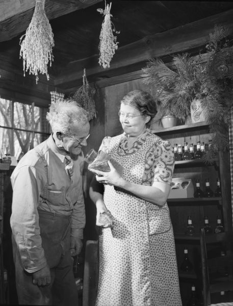Elderly couple standing in a room with dried herbs hanging from the rafters, and bottles on a shelf in the background. The man is smelling dried herbs in a jar held by the woman.