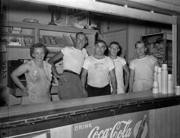 Group portrait of four snack bar employees, one woman and four men,  standing behind a counter. Behind them are shelves with a display of candy in boxes. Stacks of cups on the counter on the right, and on a shelf behind is a large stack of potato chip bags. Signs advertising "Coca Cola" and "Dad's Root Beer" are on the wall below the counter and above the shelf of candy.