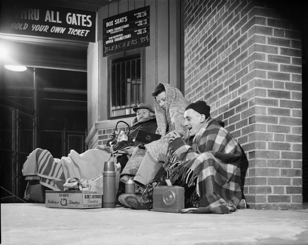 Three men wrapped up in blankets waiting at a stadium box office for tickets to go on sale. The man in the middle is reading a book. The man on the right has a thermos and a radio. The man on the left is sitting in a chair holding a lantern.