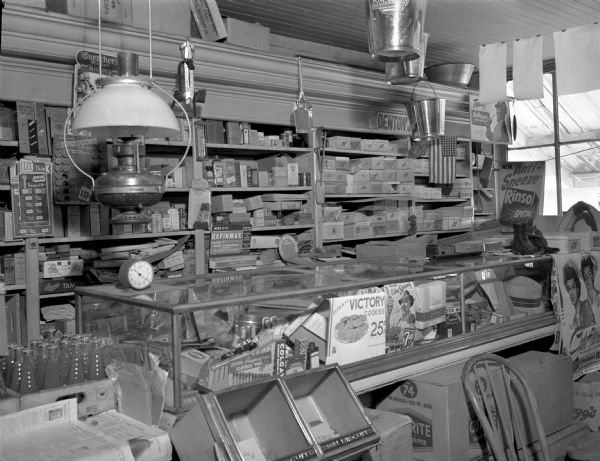 Interior view of Smith's Store, featuring a glass topped case filled with merchandise. Pails and a large light fixture hang from the ceiling, and boxes of merchandise are along the back wall on shelves. There is a window on the right looking out to the street.