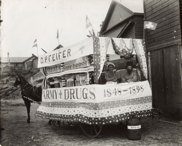 View of a horse-drawn float decorated with banners and flags and rows of pharmaceuticals on shelves. Two costumed children are posing in the back. A large container marked "Rose Oil" is on the ground behind the float. Identified as the Charles Pfeifer Float in a Spanish-American War Parade.